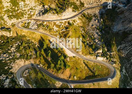 Route sinueuse serpente dans les hautes terres parmi les forêts et les collines au coucher du soleil. Les voitures roulent sur l'autoroute serpentine dans les montagnes pittoresques dans la vue aérienne ensoleillée du soir Banque D'Images