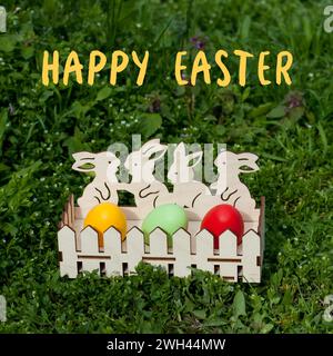 Carte de voeux avec texte Happy Easter en anglais. Coquetier en bois mignon avec des oeufs de Pâques peints debout sur l'herbe verte dans le jardin de printemps. Traditi de Pâques Banque D'Images