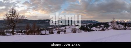 Petit village alpin et hiver montagne enneigée au premier lever du soleil autour, Voronenko, Carpates, Ukraine. Tracer sur un chemin fraîchement trodden à travers g Banque D'Images