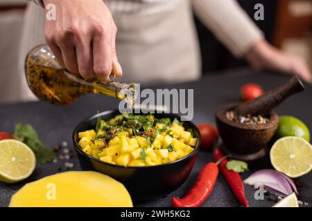 Cuisson de la salsa à la mangue - femme chef versant de l'huile d'olive de cuisson sur des ingrédients hachés tranchés dans un bol en céramique noire Banque D'Images