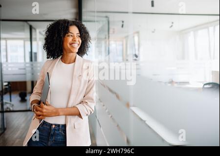 Charmante jeune femme d'affaires afro-américaine souriante dans des poutres décontractées intelligentes avec un sourire amical, son ordinateur portable à la main, mettant en valeur une atmosphère de travail accueillante. Employée de bureau transportant un ordinateur portable Banque D'Images