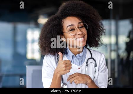 Une femme médecin avec des cheveux noirs et des lunettes est joyeusement souriante, montrant un geste du pouce vers le haut, lors d'un événement de soins de la vue. Banque D'Images