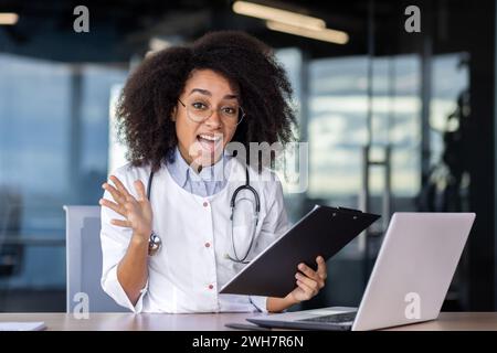 Une femme médecin, vêtue d'un manteau blanc, est assise à un bureau dans une pièce, travaillant sur un ordinateur portable et un presse-papiers avec un sourire sur le visage. Banque D'Images
