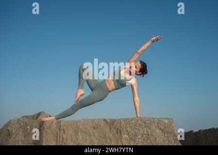 femme en forme faisant une pose de yoga de planche latérale sur les rochers, fond de ciel bleu Banque D'Images