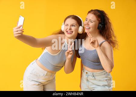 Soeurs rousses heureuses dans des écouteurs prenant selfie sur fond jaune Banque D'Images