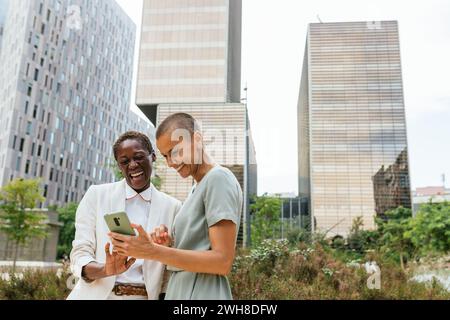 deux femmes d'affaires dans un parc devant des bâtiments d'affaires contemplant un smartphone Banque D'Images