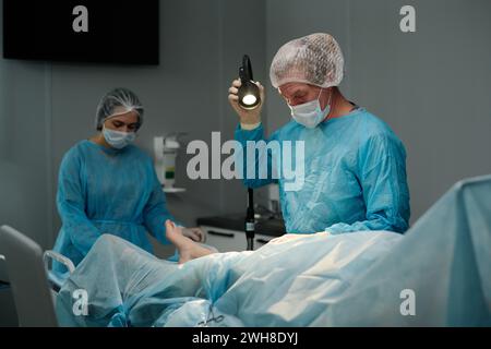 Chirurgien masculin ajustant la lampe avant l'opération chirurgicale tout en se tenant debout près de la table d'opération avec le patient recouvert d'une serviette Banque D'Images
