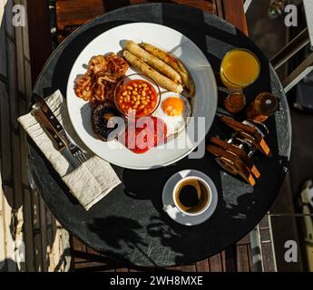 Petit déjeuner anglais complet avec œufs frits, saucisses, bacon, haricots, toasts et café servis sur table ronde le matin ensoleillé d'été Banque D'Images