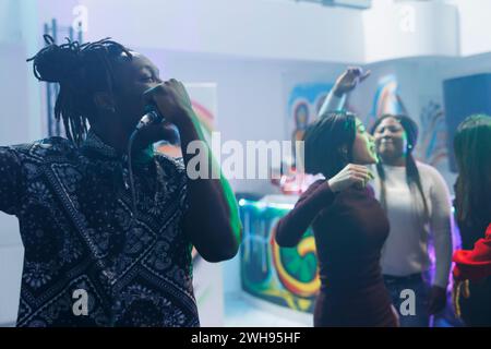 Homme afro-américain chantant dans le micro tout en dansant avec des amis dans une boîte de nuit bondée. Jeune clubber tenant le micro tout en faisant la fête à la discothèque Banque D'Images