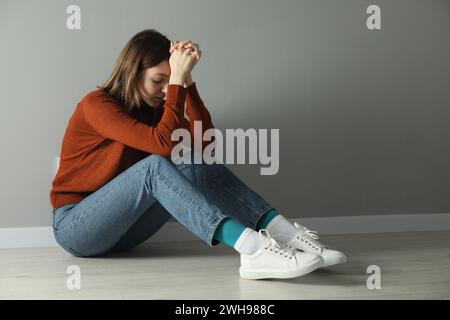 Triste jeune femme assise sur le sol près du mur gris à l'intérieur Banque D'Images