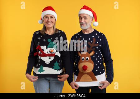 Heureux couple senior dans les chapeaux de Père Noël montrant des chandails de Noël sur fond orange Banque D'Images