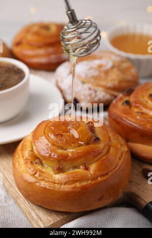 Verser du miel sur de délicieux rouleaux avec des raisins secs à table, gros plan. Petits pains sucrés Banque D'Images