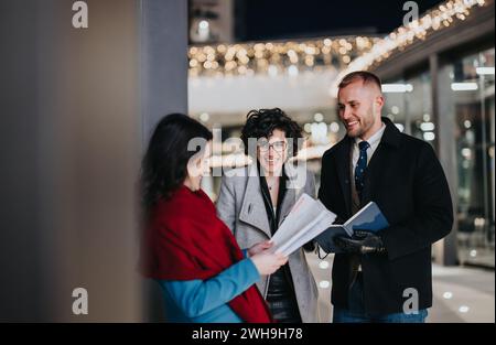 De jeunes associés joyeux s'engagent dans une conversation éclairée avec des documents dans une rue urbaine ornée de lumières festives. Banque D'Images