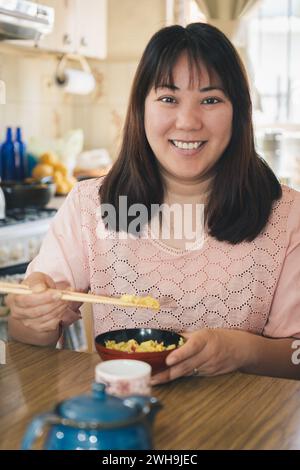 Portrait vertical d'une femme asiatique assise à la maison mangeant du riz avec des baguettes regarde et sourit à la caméra. Banque D'Images