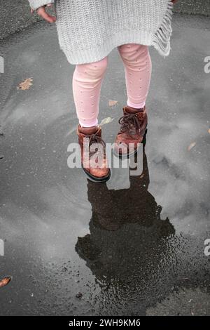 Fille en collants roses pull gris et chaussures marron auto réflexion dans la flaque de pluie avec ondulations sur la route Banque D'Images