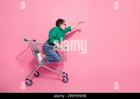 Photo pleine taille de personne positive porter un pantalon en denim soulevant le tour de poing à l'espace vide sur le chariot isolé sur fond de couleur rose Banque D'Images