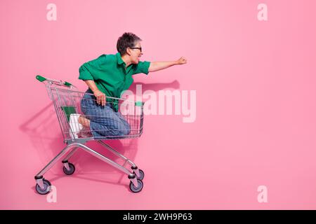 Photo du corps entier de la femme funky porter une chemise verte chevauchant le panier dirigeant poing dans l'espace vide isolé sur fond de couleur rose Banque D'Images