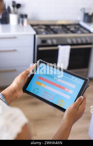 Jeune femme biraciale utilise une application de maison intelligente sur une tablette dans une cuisine moderne Banque D'Images