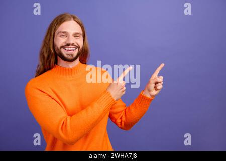 Portrait de jeune homme beau blond à cheveux longs portant orange pull tendance nouveauté doigt direct isolé sur fond de couleur violette Banque D'Images
