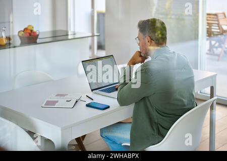Homme mûr regardant l'ordinateur assis à la table de cuisine hybride travaillant à la maison. Banque D'Images