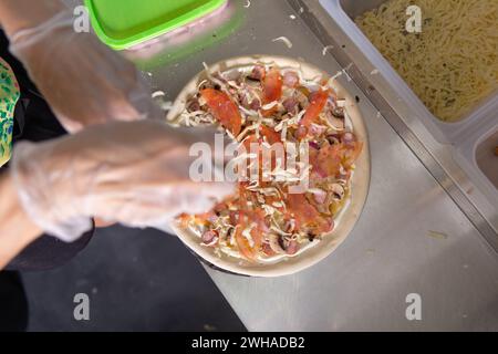 Le chef prépare une pizza italienne classique avec des saucisses, des champignons et des tomates. Le cuisinier saupoudre la pizza de fromage râpé. Vue de dessus. Banque D'Images