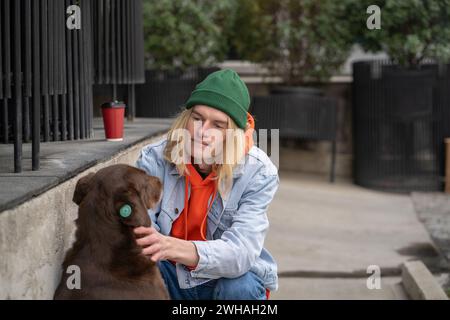 Gentil gars élégant au cœur chaleureux dans une tenue à la mode assis sur des hunches, caressant chien de rue avec amour Banque D'Images