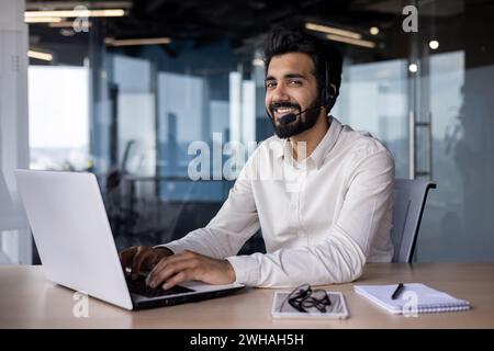 Joyeux homme d'affaires indien portant un casque travaillant à son ordinateur portable dans un bureau moderne. Jeune entrepreneur au travail. Banque D'Images