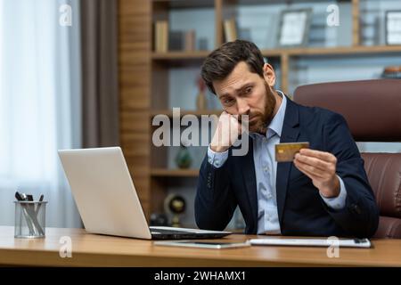 Homme adulte pensif en tenue d'affaires travaillant à la maison, tenant une carte de crédit et profondément concentré sur l'écran d'ordinateur portable. Banque D'Images