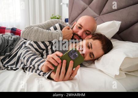 Couple gay heureux utilisant le téléphone ensemble dans le lit. Banque D'Images