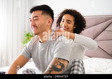 Femme latine massant les épaules de son petit ami asiatique sur le lit. Banque D'Images