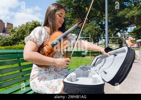 Jeune femme latine violoniste vénézuélienne artiste de rue, assise sur un banc en bois dans le parc, se préparant à jouer et à travailler. concept de personnes, espace de copie. Banque D'Images