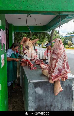 Une tête de porc et de la viande sur des hameçons à viande dans une boucherie en plein air à Bonao, République dominicaine. Le boucher découpe la carcasse en arrière-plan en deux Banque D'Images