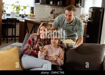 Une famille de trois est confortablement nichée sur un canapé, leurs visages reflétant l'excitation et l'attention comme ils partagent un bol de pop-corn pendant un susp Banque D'Images