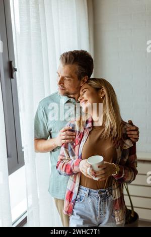 Dans une pièce légèrement éclairée, un homme embrasse amoureusement une femme par derrière alors qu'ils regardent tous les deux par la fenêtre, partageant ensemble un moment calme et intime Banque D'Images