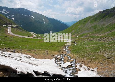 Une route sinueuse serpente à travers une vallée verdoyante avec des champs de neige, Capra River, route de montagne, Transfogarasan High Road, Transfagarasan Banque D'Images