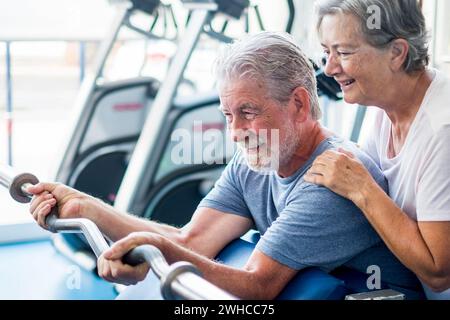 beau couple de deux personnes âgées à la salle de gym faire des exercices avec une forte lumière à l'arrière-plan - femme embrassant son mari tenant un barr - mode de vie actif Banque D'Images