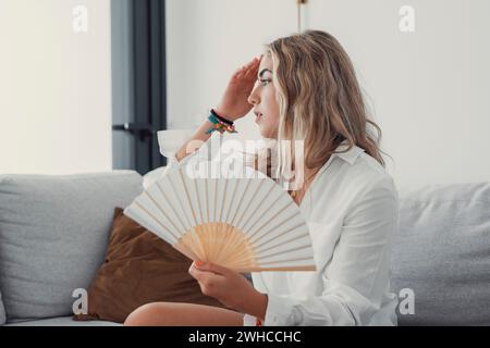 Femme surchauffée assise sur le canapé dans le salon à la chaude journée de temps d'été ressentant l'inconfort souffre de chaleur agitant le ventilateur blanc pour se refroidir, fille transpirant logement sans concept de climatiseur Banque D'Images