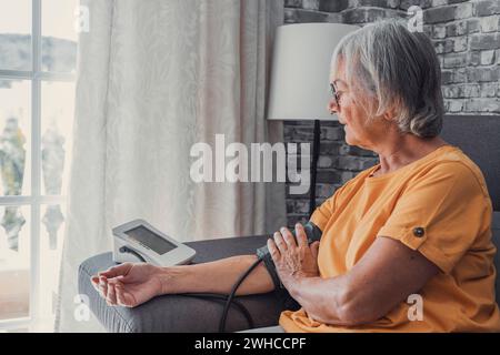 La femme aînée principale mesure le test d'hypertension artérielle basse en utilisant le compteur électronique numérique médical assis sur le canapé à la maison, prévention de l'athérosclérose des maladies cardiaques, concept d'hypertension d'hypotension Banque D'Images