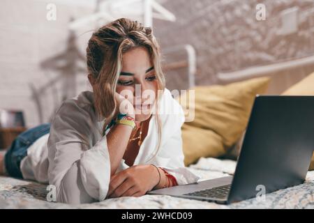 Gros plan d'une jeune femme ennuyée utilisant un ordinateur portable à l'intérieur à la maison couché sur le lit surfant sur le net. Adolescente fatiguée du millénaire étudiant en ligne. Banque D'Images