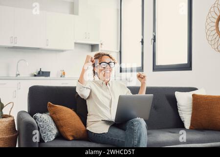 Femme excitée regardant l'écran d'ordinateur portable, criant de joie, surprise par le message avec de bonnes nouvelles, jeune femme ravie assise sur le canapé à la maison, en utilisant l'ordinateur, célébrant la victoire ou le succès en ligne Banque D'Images