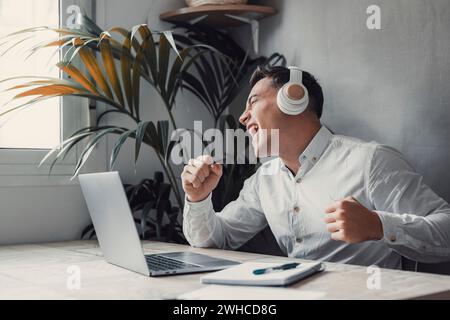 Homme d'affaires heureux portant des écouteurs chantant la chanson sur le lieu de travail, employé drôle ou freelance à l'aide d'un ordinateur portable, appréciant la piste préférée, écoutant de la musique Banque D'Images