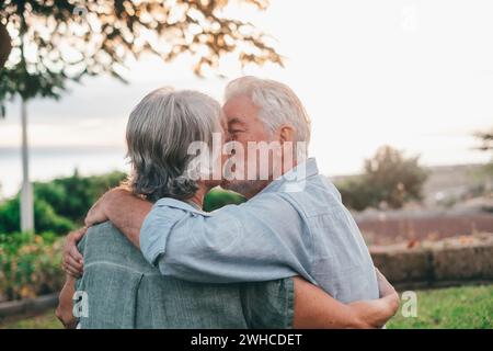 Portrait gros plan portrait heureux aux cheveux gris femme d'âge moyen blottie à son mari plus âgé souriant, profitant d'un moment tendre au parc. Lier aimant vieux couple de famille embrassant, regardant le coucher du soleil. Banque D'Images