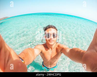 Portrait d'un jeune homme heureux prenant un selfie de lui sur la plage dans une eau turquoise bleue ayant du plaisir et appréciant seul des vacances à l'extérieur. Banque D'Images