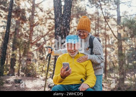 Portrait, gros plan de couple de personnes âgées mignonnes utilisant le téléphone ensemble dans la forêt avec des arbres autour d'eux. Utilisation de téléphone portable mobile à l'extérieur s'amuser et profiter. Banque D'Images