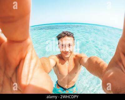 Portrait d'un jeune homme heureux prenant un selfie de lui sur la plage dans une eau turquoise bleue ayant du plaisir et appréciant seul des vacances à l'extérieur. Banque D'Images