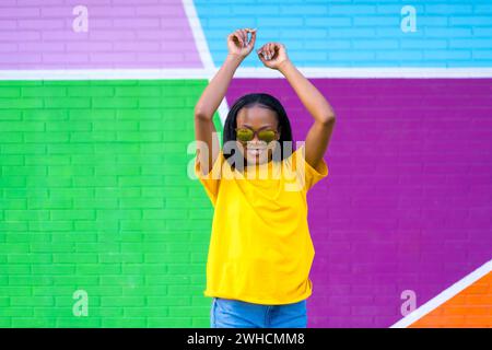 Portrait frontal d'une femme africaine avec des lunettes de soleil dansant à côté d'un mur multicolore Banque D'Images