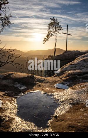 Paysage photographié sur un rocher de grès dans la forêt. Ambiance matinale au lever du soleil à un point de vue. Un petit arbre et une croix sommitale se dressent sur le Rötzenfels dans la forêt du Palatinat Banque D'Images