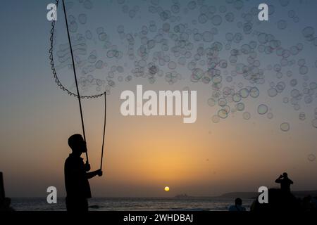 Un homme en silhouette fait beaucoup de bulles de savon avec un bâton de bulle de savon, ils flottent dans le ciel orange au coucher du soleil, certaines personnes en silhouette regardent dessus Banque D'Images