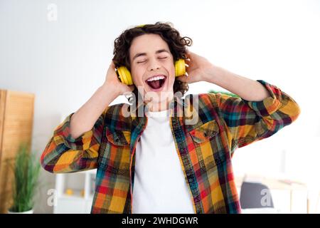 Photo de bonne humeur insouciante gars habillé chemise à carreaux écoutant des écouteurs de musique chantant à l'intérieur maison chambre à la maison Banque D'Images