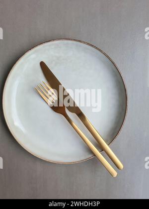 Assiette en céramique de couleur naturelle avec rebord marron et couteau de couleur or avec fourchette sur fond gris, assiette avec couverts Banque D'Images